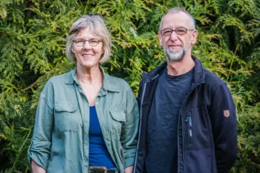 Man en vrouw poseren lachend voor een groene coniferenhaag.